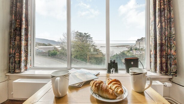 A breakfast setup with croissants on a plate, two white mugs, and a newspaper on a table by a window with a view of a coastal town. - 2 bedroom accommodation in Barmouth (Barmouth)