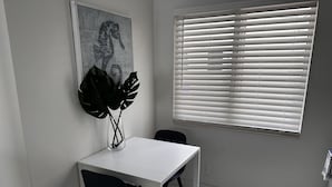 Un petit coin repas avec une table et des chaises blanches, une grande plante dans un vase, un tableau représentant un hippocampe accroché au mur et une fenêtre avec des stores.
