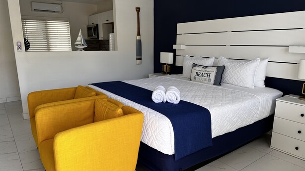 Une chambre d’hôtel moderne avec un grand lit, un fauteuil jaune et une vue sur la cuisine et l’espace de vie.
