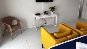 Une chambre d’hôtel avec un lit, un téléviseur fixé au mur, un bureau agrémenté d’une plante, une chaise en osier et un fauteuil jaune.