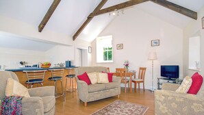 A spacious living area with a kitchen in the background, wooden beams on the ceiling, and a dining table with chairs. - Lower Bushford Cottage (Hasguard, near Dale)