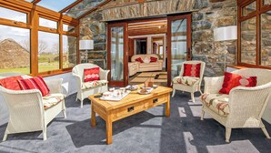 Ein geräumiger Raum mit hohen Decken, Steinwänden und großen Fenstern. Es gibt Korbstühle mit roten Kissen, einen Holztisch und eine Couch mit roten und weißen Kissen.