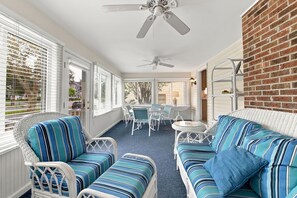 Une terrasse ensoleillée, meublée de mobilier en osier, avec un mur en briques et un ventilateur de plafond.