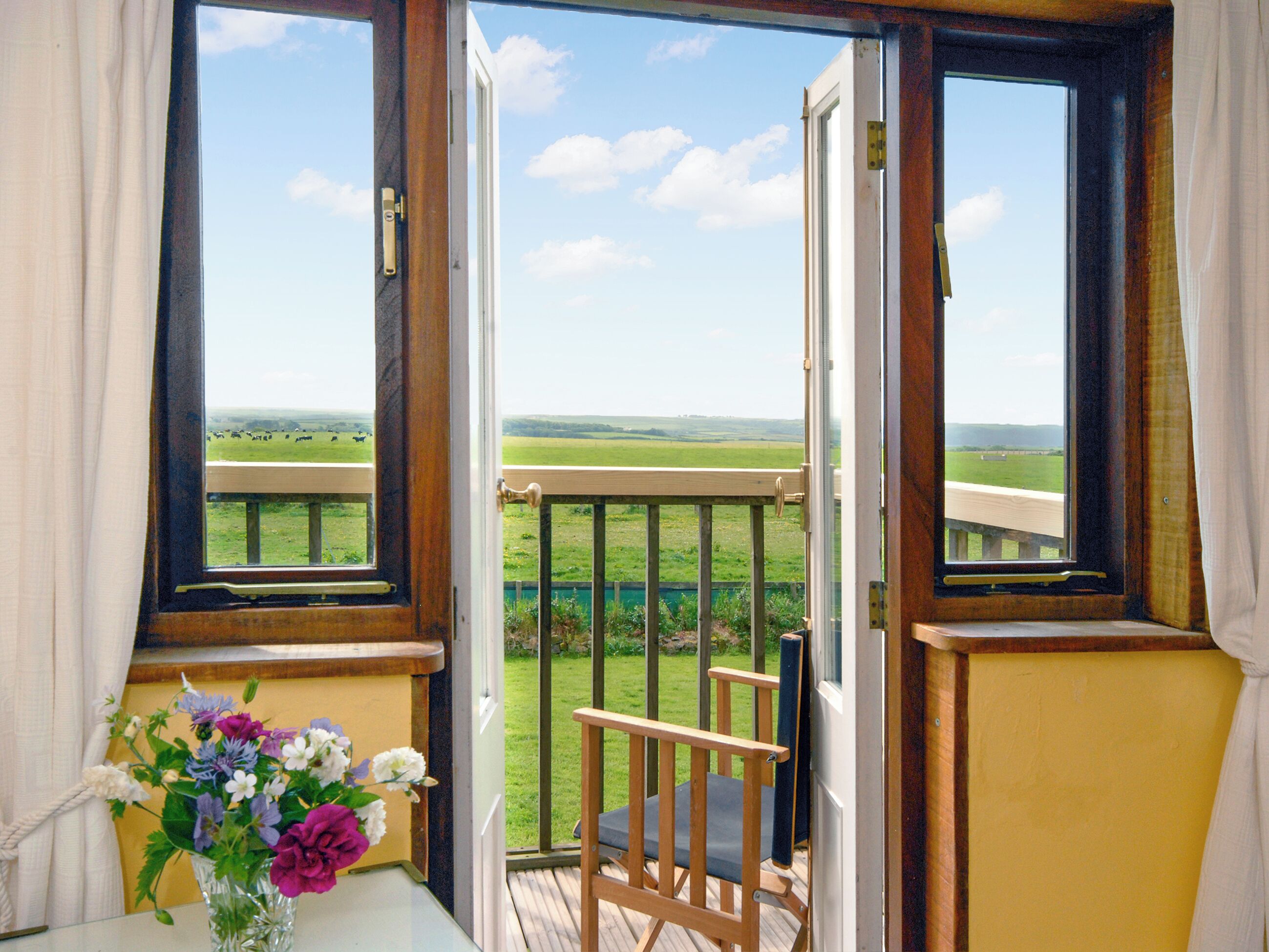 A room with a wooden chair, a table with a vase of flowers, and a view of a green field through open windows.