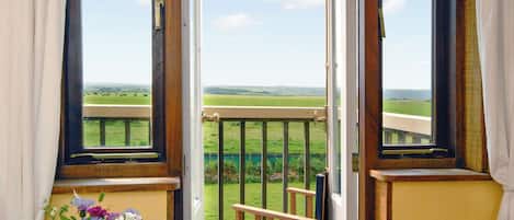 A room with a wooden chair, a table with a vase of flowers, and a view of a green field through open windows.