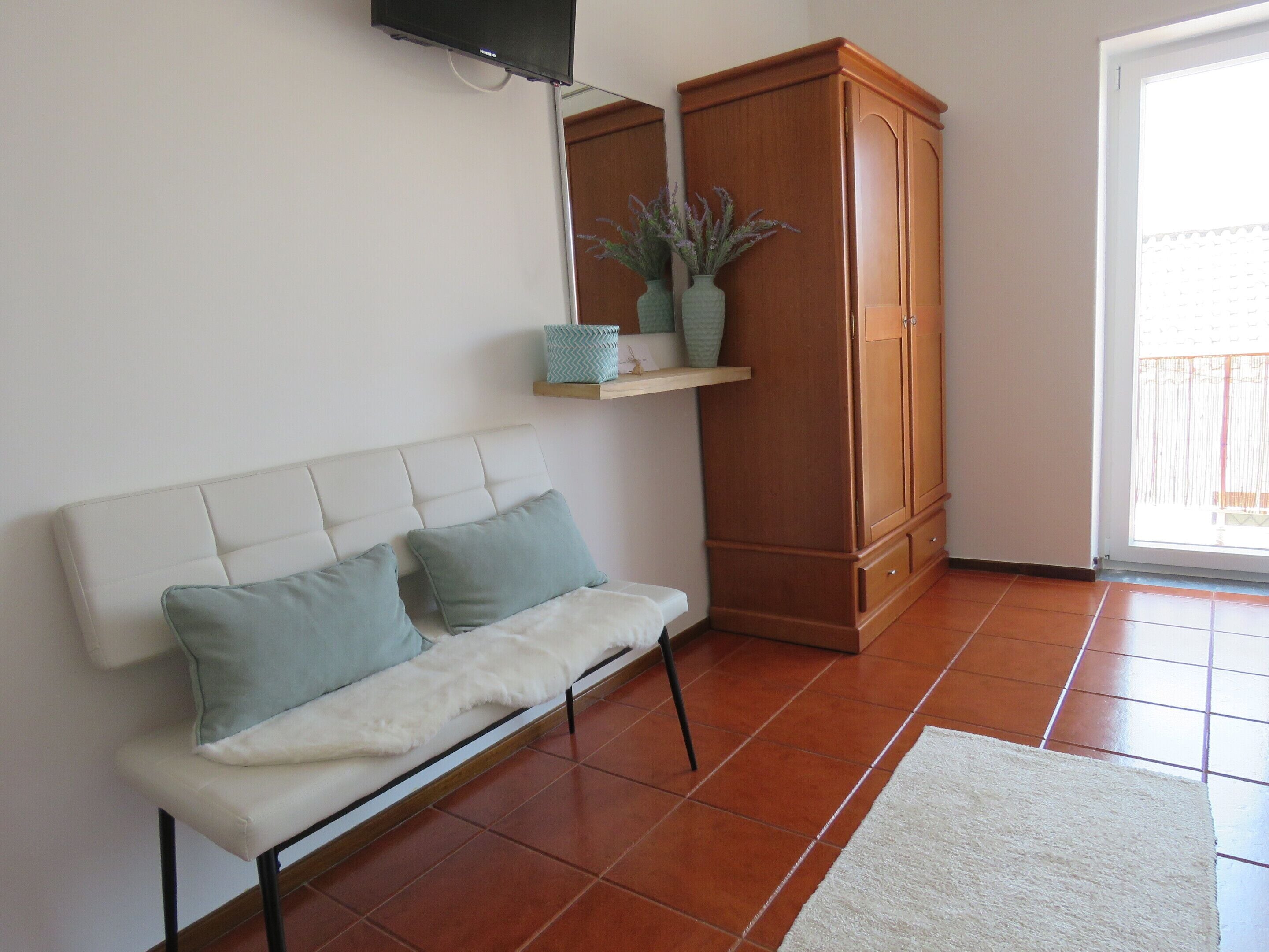 A living room with a white sofa, two light blue cushions, a white furry throw, a wooden cabinet, and a small shelf with decorative items.