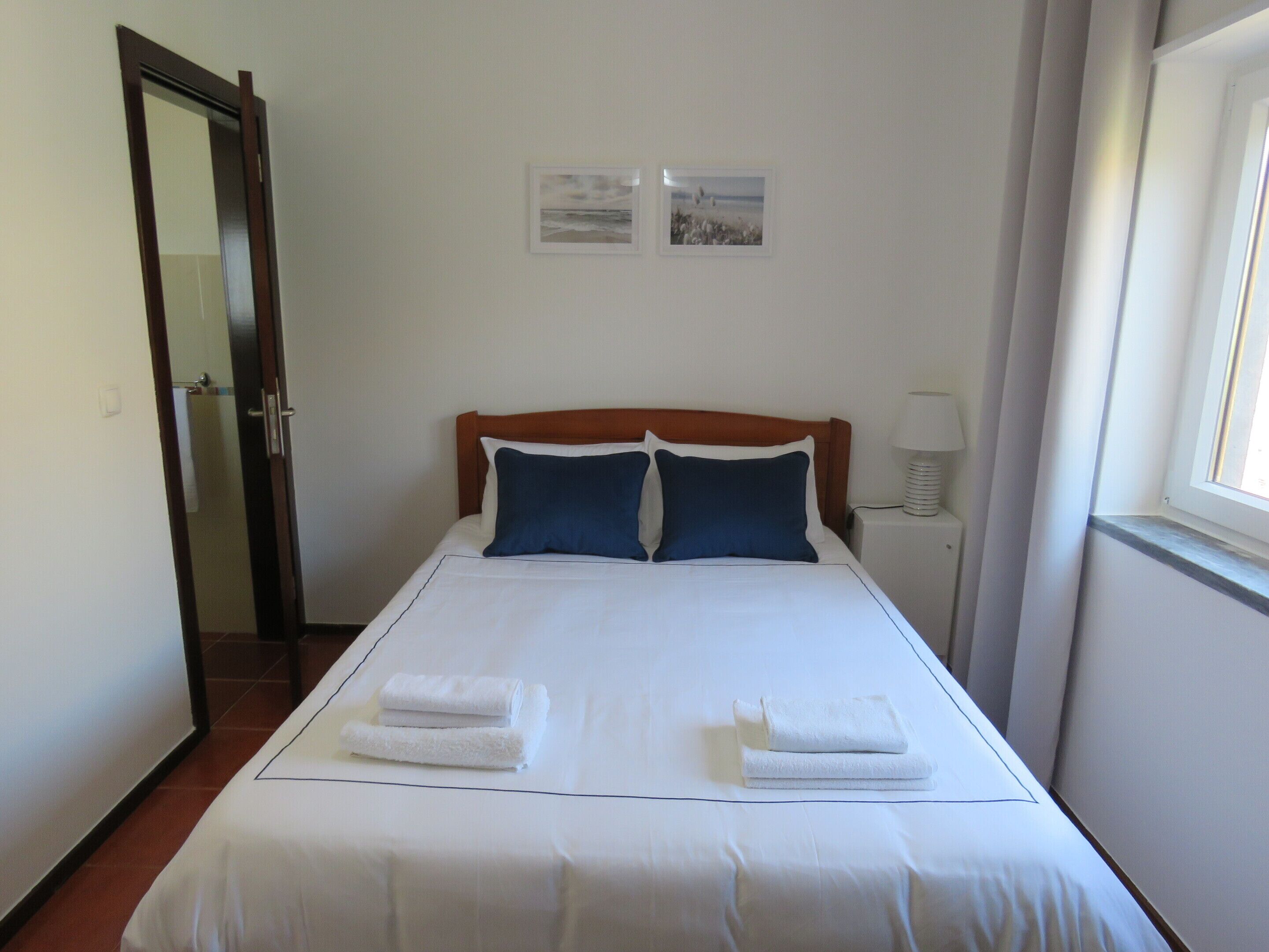 A neatly made bed with white linens and two blue pillows, a bedside lamp, and two framed pictures on the wall.