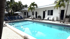Una piscina rettangolare con indicatore di profondità di 1,8 metri, circondata da sdraio e palme, con un edificio bianco sullo sfondo.