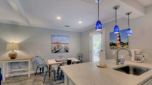 Une cuisine moderne dotée d’un plan de travail en granit, d’un évier en acier inoxydable et de luminaires suspendus. On y trouve également un coin repas avec une table et des chaises, ainsi qu’un tableau accroché au mur.