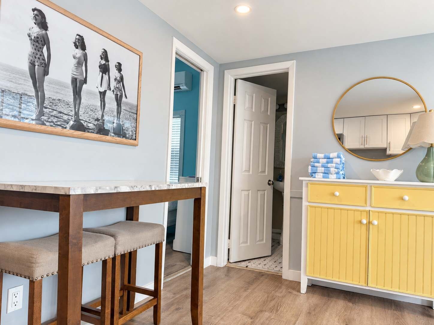 Une pièce avec une table en bois et des tabourets de bar, un meuble jaune avec des tiroirs, une photographie encadrée accrochée au mur et une porte donnant sur une autre pièce.
