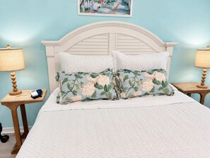 Una cama con colcha blanca y dos almohadas, una con estampado floral. Hay dos mesitas de noche de madera con lámparas, y un cuadro en la pared.
