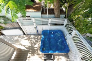 Un jacuzzi en un área de terraza de madera rodeado por una valla blanca, con árboles y un jardín al fondo.