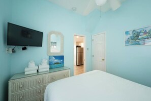 A bedroom with a bed, a dresser, a television, a mirror, and a door leading to a kitchen.