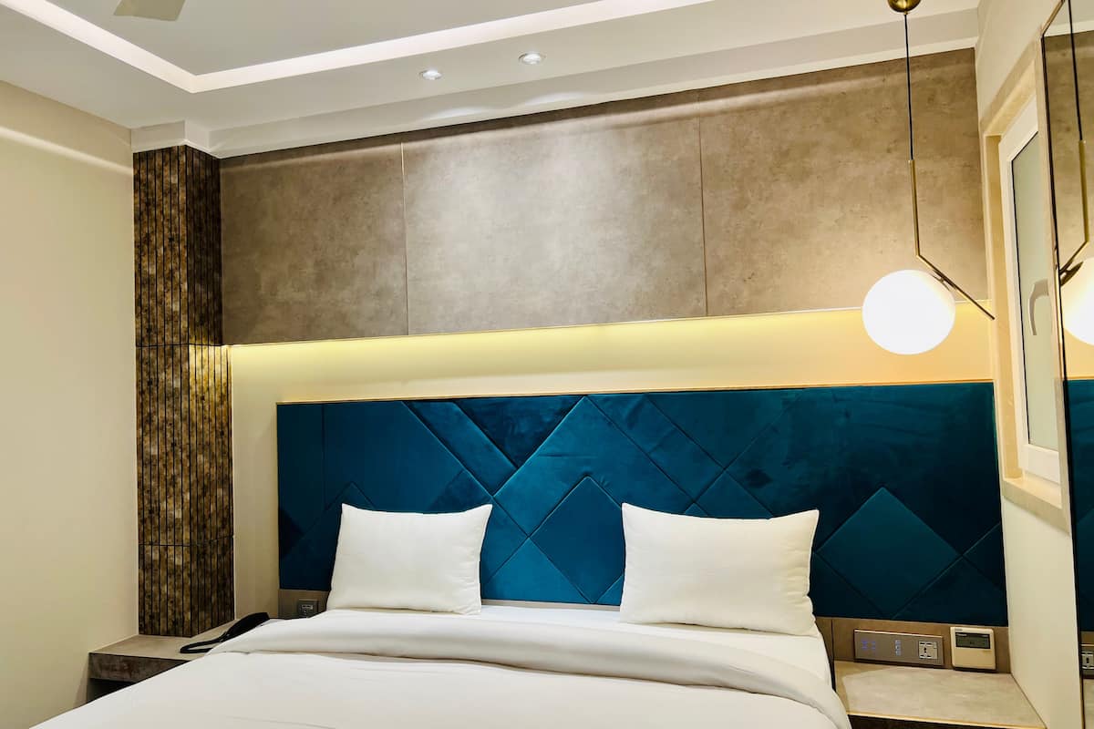 A modern hotel room with a large bed, a headboard with a geometric pattern, a bedside table with a phone and a control panel, and a ceiling light fixture.