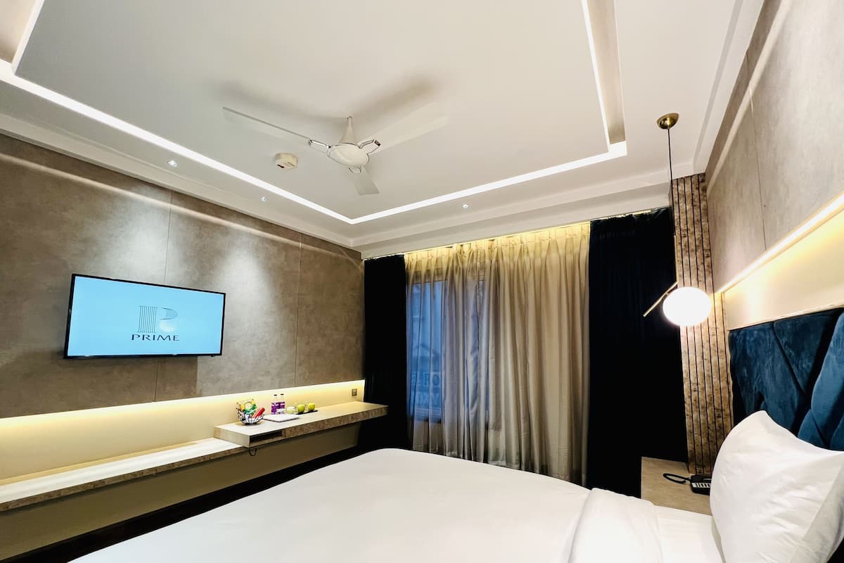 A modern hotel room with a flat-screen TV displaying the 'PRIME' logo, a ceiling fan, a bedside table with a lamp, and a large bed with white linens.