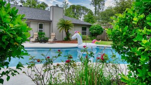 A swimming pool with a unicorn float, surrounded by lush greenery and a house with a patio.