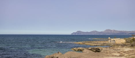 A coastal landscape with rocky terrain, a small building, and mountains in the distance.