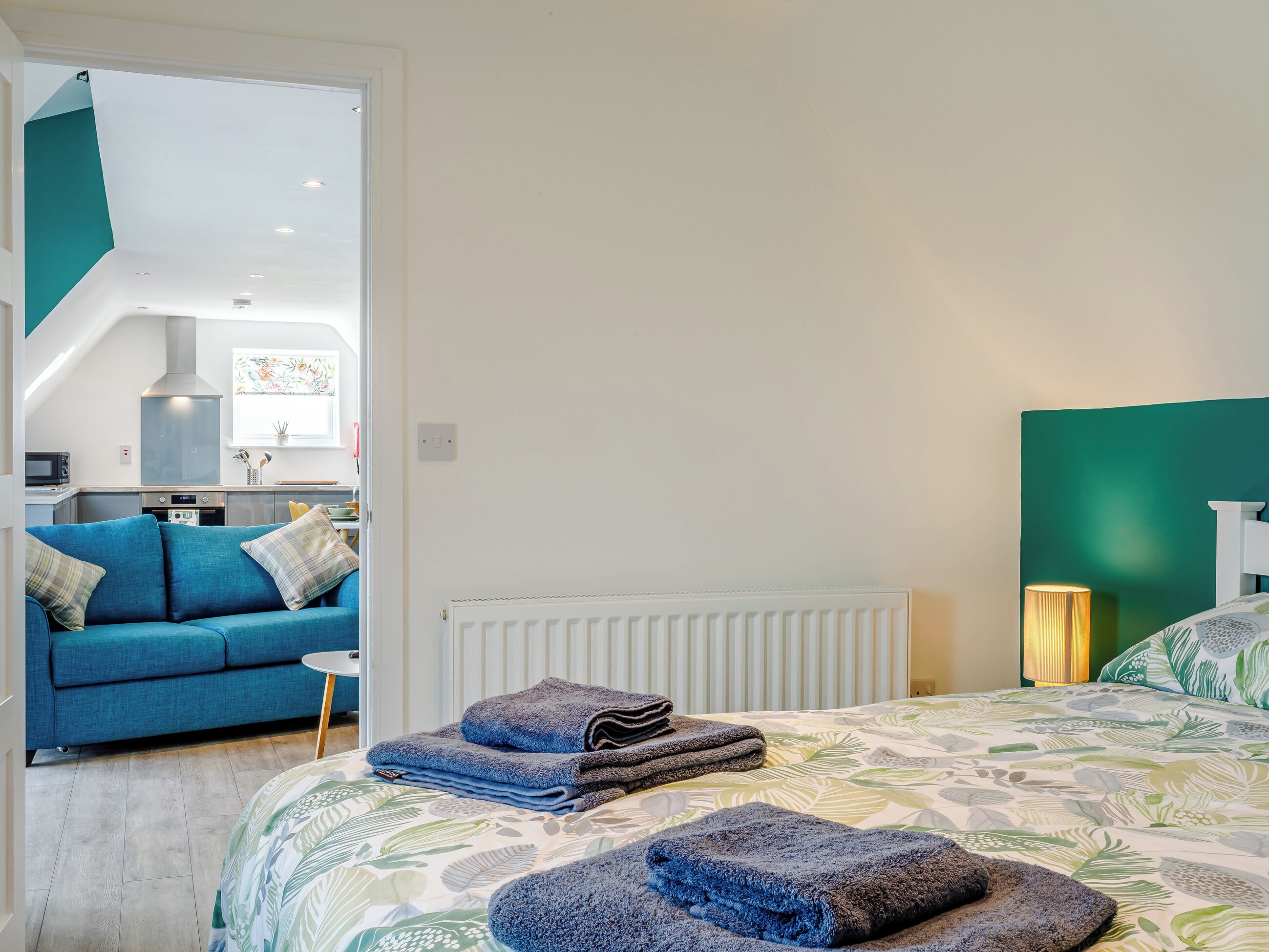 A bedroom with a bed, a radiator, a bedside lamp, a blue sofa, and a view into the kitchen.