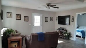 A living room with a brown sofa, a ceiling fan, a TV mounted on the wall, and a door leading to another room.
