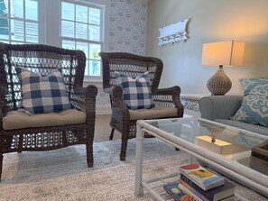 Une pièce comprenant deux fauteuils en osier avec des coussins à carreaux, une table basse en verre, un canapé gris avec des coussins à motifs, une lampe fixée au mur et une bibliothèque remplie de livres.