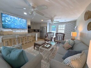 A living room with a sofa, armchairs, a coffee table, and a TV. There are two ceiling fans, a kitchen area with a refrigerator, and a dining bar with stools. - Duval Vibes • Pool • 10 Guests (Key West)