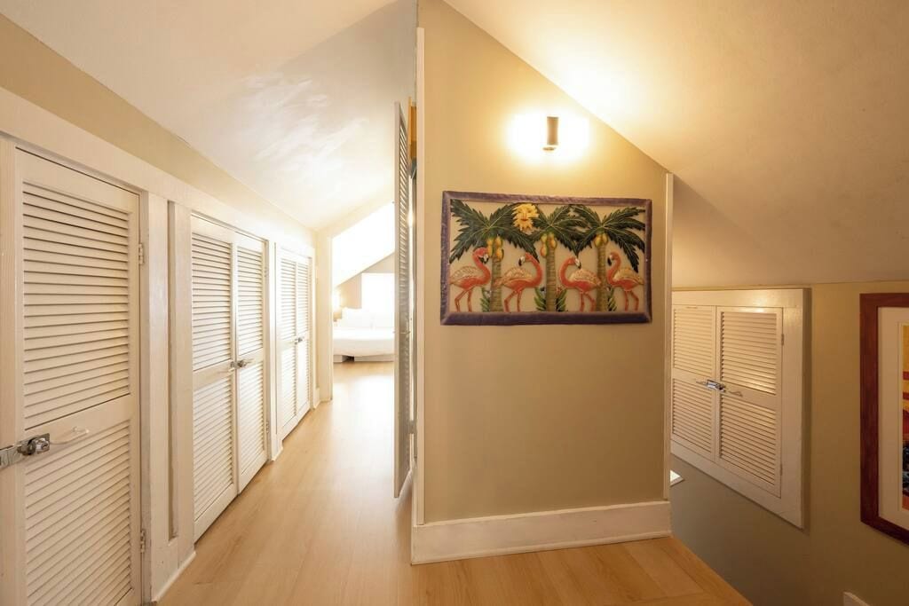 A hallway with closed louvered doors, a wall-mounted light fixture, and a framed tropical-themed artwork.