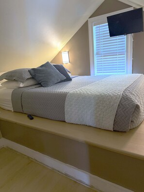 Une chambre à coucher avec un lit, des oreillers, une couverture, une télécommande, un téléviseur fixé au mur et une fenêtre avec des stores.