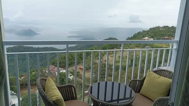Een balkon met rieten stoelen en een tafel, met uitzicht op een kustlandschap met heuvels en een waterpartij.