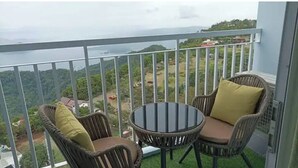 Een balkon met rieten stoelen en een tafeltje, met uitzicht op een fraai landschap met begroeiing en gebouwen in de verte.