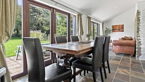 A dining area with a wooden table and chairs, large windows with curtains, a stone fireplace, and a view of the garden. - Spacious Teifi Valley barn for holidays near the Cardigan coast. (Cenarth)