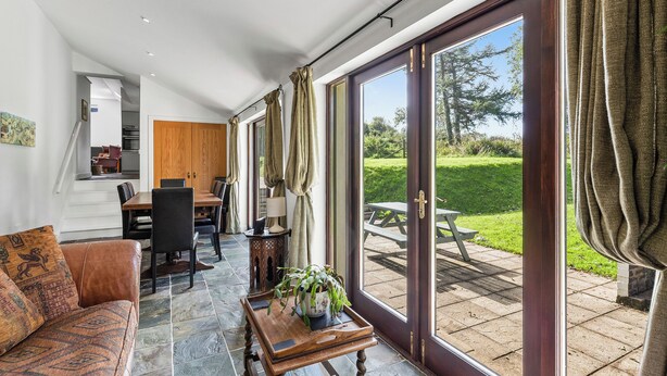 A spacious living area with a sofa, dining table, and chairs, a wooden coffee table with a plant, and large sliding glass doors leading to an outdoor patio with a picnic table. - Spacious Teifi Valley barn for holidays near the Cardigan coast. (Cenarth)