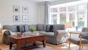Ein Wohnzimmer mit grauem Sofa, einem Holztisch und einem blauen Sessel. An der Wand befinden sich gerahmte Bilder, ein Fenster mit Blick auf Bäume und eine Lampe auf einem Beistelltisch.