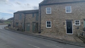 A two-story stone building with a black door and windows, located on a street with a curb and a drainage grate. - White Lea is a beautiful Dales Cottage recently transformed into a fantastic holiday home. (Richmond)