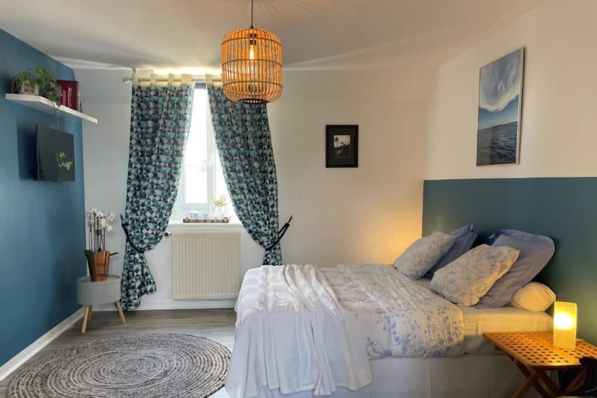 Un dormitorio con cama, mesita de noche con lámpara, ventana con cortinas y estante en la pared con plantas.