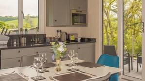 A modern kitchen with a built-in oven and microwave, a dining area with a table set for four, and a view of a green landscape through large windows.