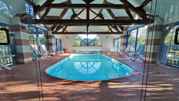An indoor swimming pool with a tiled floor, wooden beams, and a view of the outdoors through large windows.