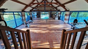 A spacious indoor area with exercise equipment, a pool visible through a glass door, and a wooden staircase leading to a balcony.