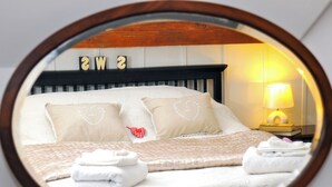 Ein ordentlich bezogenes Bett mit weißen Leinen und herzförmigen Kissen, eine Nachttischlampe und ein Spiegel, der die Raumgestaltung widerspiegelt.