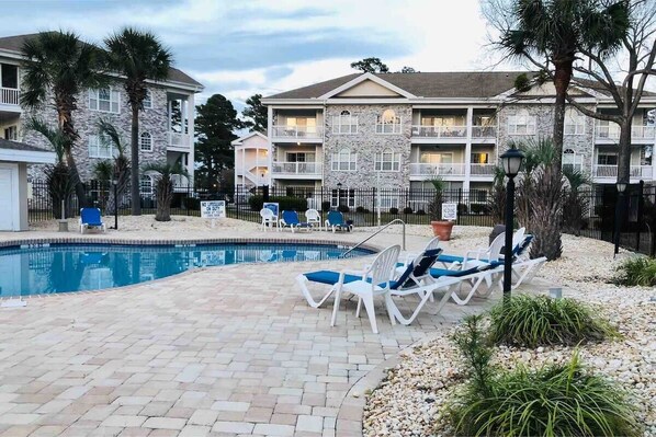 Une zone de piscine avec des chaises longues et, au fond, un bâtiment doté de nombreux balcons.