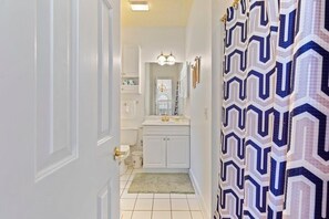 Un couloir menant à une salle de bain dotée d’un rideau de douche à motifs, de meubles blancs et d’un lustre.