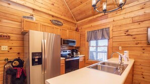 A wooden cabin kitchen with a stainless steel refrigerator, microwave, and stove. There is a sink, a window with blinds, and a teddy bear hanging on the wall. - 3 Kings / Deck / Fireplace / 100Mbps / Games Games/ Parking/ Dollywood 5mi (Sevierville)