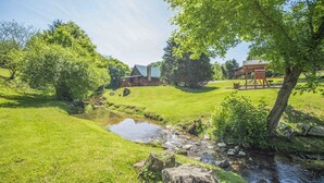 A rustic cabin with a large porch, surrounded by lush green grass and a small stream. - 3 Kings / Deck / Fireplace / 100Mbps / Games Games/ Parking/ Dollywood 5mi (Sevierville)