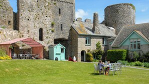 A castle with stone walls and a round tower, a small blue building, and a grassy area with tables and chairs. - 4 bedroom accommodation in Manorbier (Manorbier)