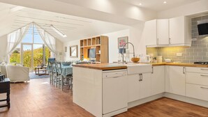 A modern kitchen with white cabinetry, a wooden countertop, and a built-in dishwasher. There is a dining area with a table and chairs, and a large window providing a view of the outdoors. - 4 bedroom accommodation in Manorbier (Manorbier)