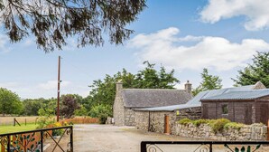 Une charmante maison de campagne en pierre, avec un toit en métal, entourée de verdure et bordée d’un chemin de gravier.