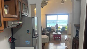Eine Küche mit Einbauherd, Kühlschrank und Holzausstattung. Es gibt eine Essecke mit Tisch und Stühlen sowie einen Blick aufs Meer durch ein großes Fenster.