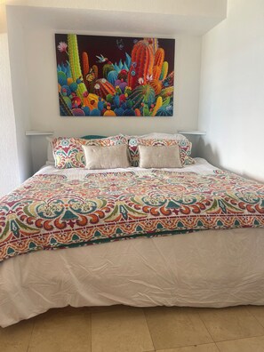 Ein Schlafzimmer mit einem farbenfrohen Bettbezug, einer weißen Daunendecke und einem lebhaften Bild von Kakteen und Blumen an der Wand.
