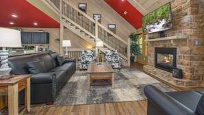 A spacious living room with a fireplace, a staircase, and a large TV mounted on a stone wall. - Smoky Mountain Plunge (Gatlinburg)