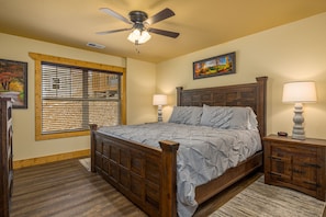 A bedroom with a wooden bed, two bedside tables with lamps, a ceiling fan, a window with blinds, and a painting on the wall.