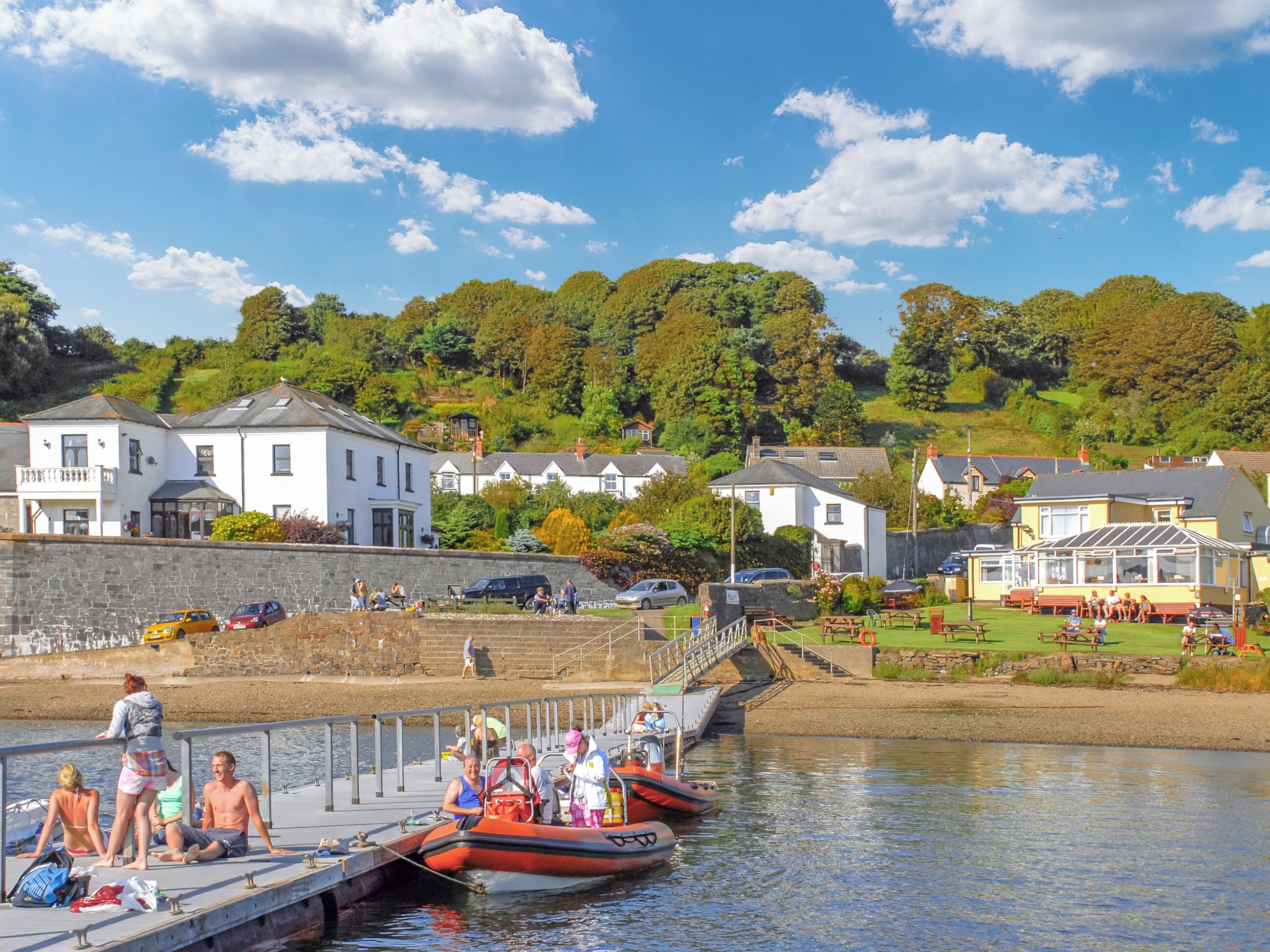A riverside pier with boats, a stone wall, and houses in the background.